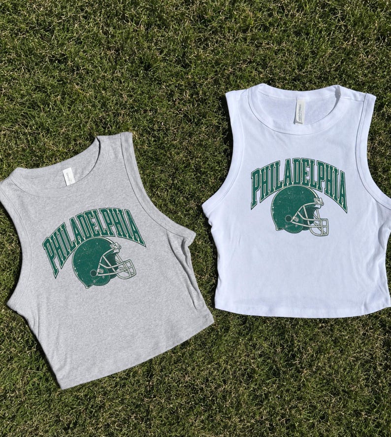 Może przedstawiać: Dwa bezrękawniki typu crop top, jeden w kolorze szarego melanżu, a drugi biały, każdy z ciemnozieloną grafiką kasku do futbolu amerykańskiego i napisem PHILADELPHIA łukowatą czcionką. Wzory mają postarzany, przetarty wygląd.