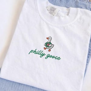 May include: A white short-sleeve t-shirt featuring an embroidered graphic of a goose wearing a green jersey and holding a brown football. Below the goose, the text philly goose is embroidered in green cursive script. The shirt has a crew neckline.
Philadelphia Football, Embroidered Crewneck Sweatshirt, T-Shirt Embroidery, Philly Goose, Silly Goose
philly goose philly goose sweatshirt philadelphia philly goose shirt retro philadelphia designs philly crewneck