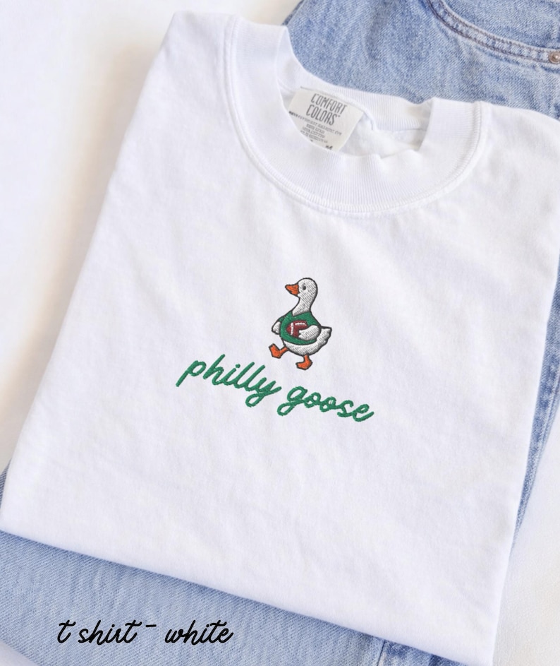 May include: A white short-sleeve t-shirt featuring an embroidered graphic of a goose wearing a green jersey and holding a brown football. Below the goose, the text philly goose is embroidered in green cursive script. The shirt has a crew neckline.
Philadelphia Football, Embroidered Crewneck Sweatshirt, T-Shirt Embroidery, Philly Goose, Silly Goose
philly goose philly goose sweatshirt philadelphia philly goose shirt retro philadelphia designs philly crewneck