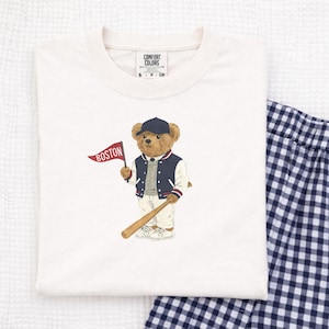 May include: Cream-colored Comfort Colors t-shirt featuring a cartoon bear wearing a baseball cap and jacket, holding a baseball bat and a flag that says "BOSTON". The shirt has a crew neck and the Comfort Colors logo.