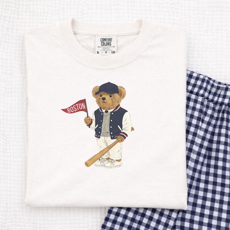 May include: Cream-colored Comfort Colors t-shirt featuring a cartoon bear wearing a baseball cap and jacket, holding a baseball bat and a flag that says "BOSTON". The shirt has a crew neck and the Comfort Colors logo.