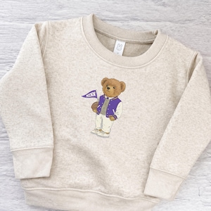 May include: A beige heathered crewneck sweatshirt featuring a central graphic of a brown teddy bear wearing a purple and white varsity jacket and white pants, holding a small purple pennant with the text MINN printed in white letters.