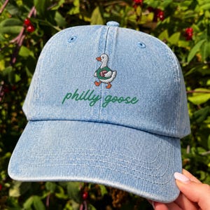 May include: A light blue denim baseball cap featuring an embroidered graphic of a white goose wearing a green jersey and holding a brown football. Below the goose, the text philly goose is embroidered in a cursive green font on the front panel.