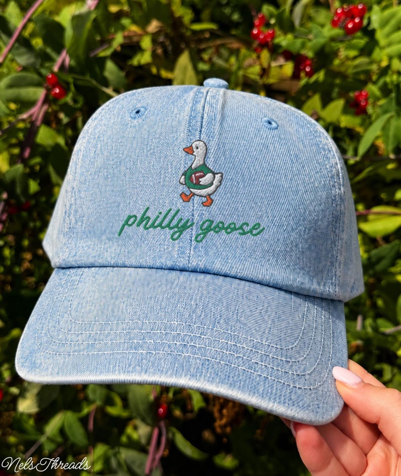 May include: A light blue denim baseball cap featuring an embroidered graphic of a white goose wearing a green jersey and holding a brown football. Below the goose, the text philly goose is embroidered in a cursive green font on the front panel.