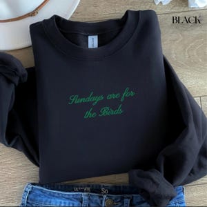 K&ouml;nnte beinhalten: Schwarzes Sweatshirt mit gr&uuml;n gesticktem Text "Sundays are for the Birds". Das Sweatshirt ist auf einer blauen Jeans gefaltet. Das Wort "BLACK" ist oben rechts aufgedruckt.