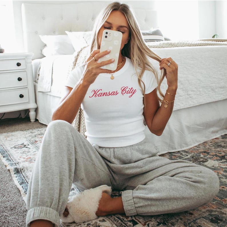 May include: White t-shirt with the text "Kansas City" in red script. The person is wearing gray sweatpants and fluffy white slippers. The person is holding a phone with a floral case.