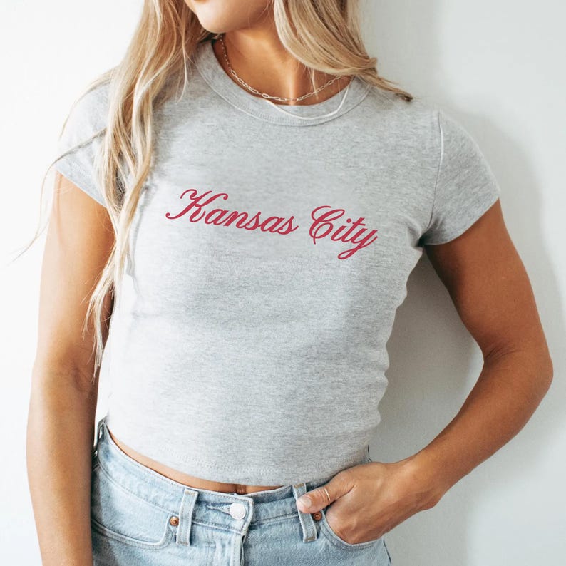 May include: A gray crop top with the text "Kansas City" in a cursive, red font. The shirt has short sleeves and is paired with blue jeans. The model is wearing a gold necklace.