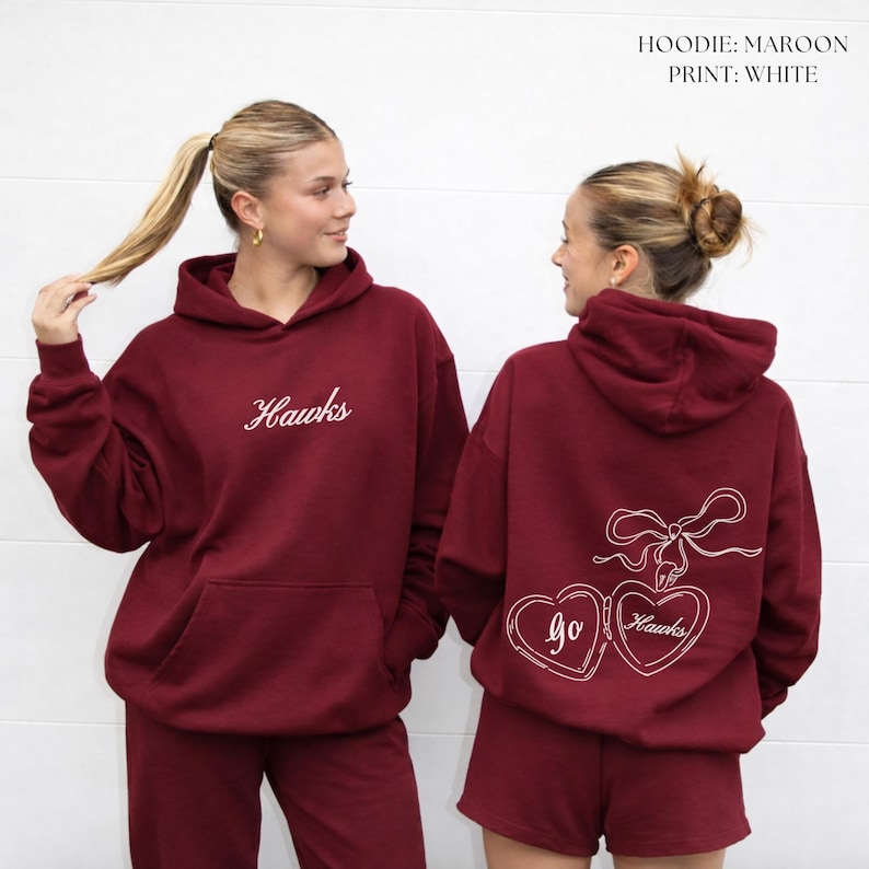 Puede incluir: Dos sudaderas con capucha color granate con texto y gr&aacute;ficos blancos. Una sudadera tiene "Hawks" bordado en la parte delantera. La otra tiene un lazo y dos medallones en forma de coraz&oacute;n con "Go Hawks" en la parte trasera. Las sudaderas se combinan con pantalones cortos y pantalones granates a juego.
