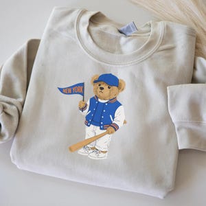 Può includere: Felpa beige chiaro con un orsetto dei cartoni animati con berretto e giacca da baseball blu, che tiene una mazza da baseball e una bandiera blu con la scritta "NEW YORK". L'orso indossa pantaloni e scarpe da ginnastica bianche.
