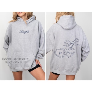 Puede incluir: Una sudadera con capucha gris claro con cord&oacute;n. La parte delantera presenta la palabra "Knights" en azul marino. La parte trasera muestra dos medallones en forma de coraz&oacute;n conectados con las palabras "go Knights" y un lazo.