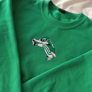 Può includere: Una felpa girocollo verde brillante con un ricamo centrale raffigurante un giocatore di football in divisa verde e bianca che esegue una posa dab. La felpa presenta un colletto a coste, maniche lunghe e una morbida fodera interna in pile.