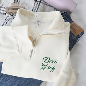 Puede incluir: Sudadera color crema con cuello con cremallera y el texto bordado "Bird Gang" en verde. La sudadera está doblada sobre unos vaqueros azules. También se ven una pinza para el pelo y un bolso.