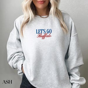May include: Ash gray sweatshirt with the text "LET'S GO Buffalo" in blue and red script. The sweatshirt is a crew neck style and has long sleeves. The word "ASH" is printed in the bottom left corner.