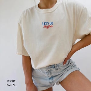 Puede incluir: Camiseta de color marfil con el texto "LET'S GO Buffalo" en escritura azul y roja. La camiseta es de corte holgado y tiene mangas cortas. Talla XL.