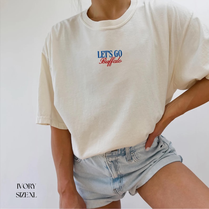 Puede incluir: Camiseta de color marfil con el texto "LET'S GO Buffalo" en escritura azul y roja. La camiseta es de corte holgado y tiene mangas cortas. Talla XL.
