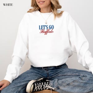 May include: White sweatshirt with the text "LET'S GO Buffalo" in blue and red. The sweatshirt is long-sleeved and has a crew neck. The model is wearing blue jeans and black and white sneakers.