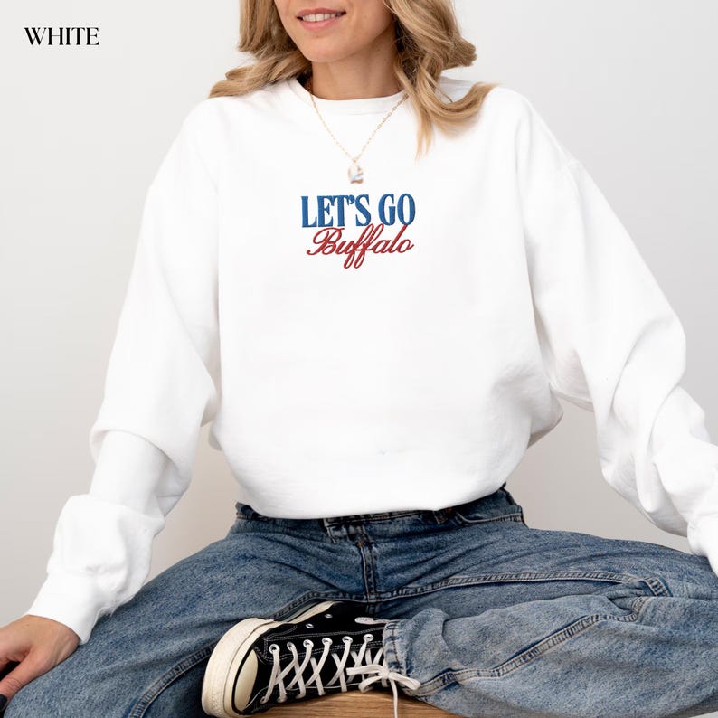 May include: White sweatshirt with the text "LET'S GO Buffalo" in blue and red. The sweatshirt is long-sleeved and has a crew neck. The model is wearing blue jeans and black and white sneakers.