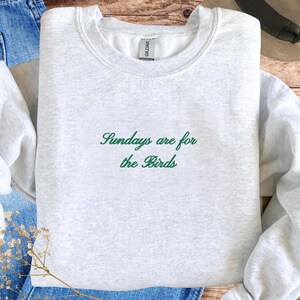 K&ouml;nnte beinhalten: Hellgraues Sweatshirt mit dem gr&uuml;nen Schriftzug "Sundays are for the Birds". Das Rundhals-Sweatshirt ist gefaltet und liegt auf einer blauen Jeans.
