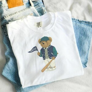May include: White t-shirt with a cartoon bear wearing a baseball cap and jacket, holding a baseball bat and a flag that says "SEATTLE". The shirt is folded on top of a pair of blue jeans.