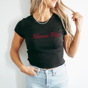 May include: Black short-sleeved t-shirt with the cursive text "Kansas City" in red. The shirt is paired with light blue denim jeans. The model is wearing a silver necklace.