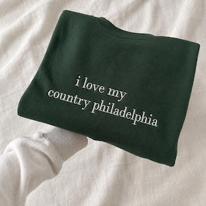 Può includere: Una felpa girocollo verde bosco scuro con il testo ricamato in bianco che recita i love my country philadelphia centrato sul davanti. Il capo presenta un classico design a maniche lunghe con scollo e polsini a coste.