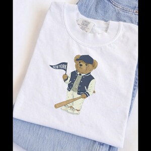 Op de afbeelding: Een wit t-shirt met een afbeelding van een bruine teddybeer die een marineblauwe baseballpet, een marineblauw met wit varsity-jack en een lichte broek draagt. De beer houdt een kleine marineblauwe vlag met de tekst NEW YORK en een honkbalknuppel vast.
