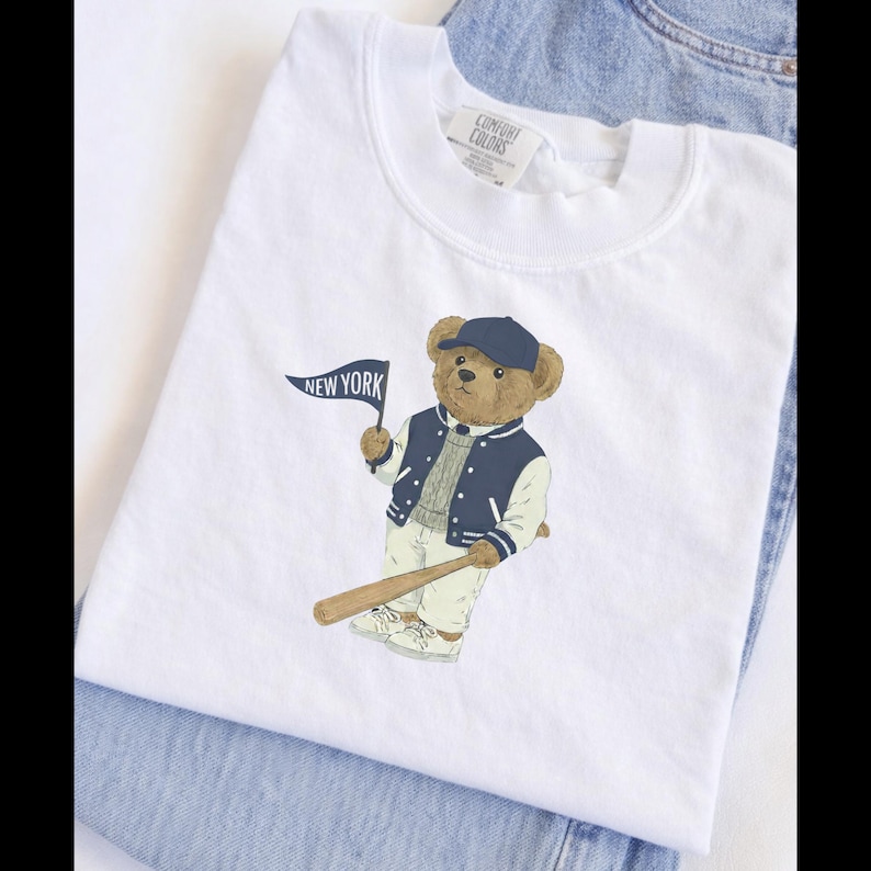 Op de afbeelding: Een wit t-shirt met een afbeelding van een bruine teddybeer die een marineblauwe baseballpet, een marineblauw met wit varsity-jack en een lichte broek draagt. De beer houdt een kleine marineblauwe vlag met de tekst NEW YORK en een honkbalknuppel vast.