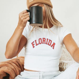 May include: White crop top with the word "FLORIDA" in orange and blue lettering. The person is wearing a brown beanie and holding a black mug. The shirt is paired with grey sweatpants.
