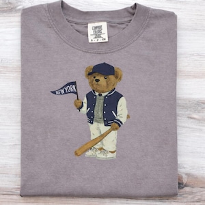 Op de afbeelding: Een grijs t-shirt met korte mouwen en een afbeelding van een bruine teddybeer met een marineblauwe baseballpet, een marineblauw-wit varsity-jack en een lichte broek. De beer houdt een blauwe wimpel met de tekst NEW YORK en een honkbalknuppel vast.