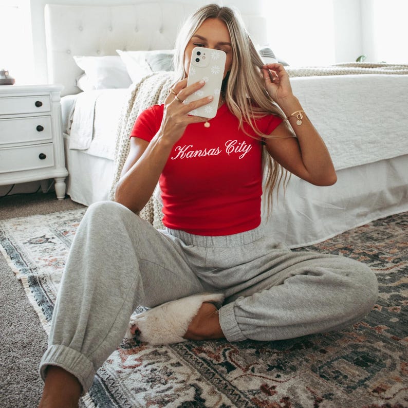 May include: A woman wearing a red crop top with the words "Kansas City" in white script. She is also wearing gray sweatpants and white fuzzy slippers. She is holding a phone with a white floral case.