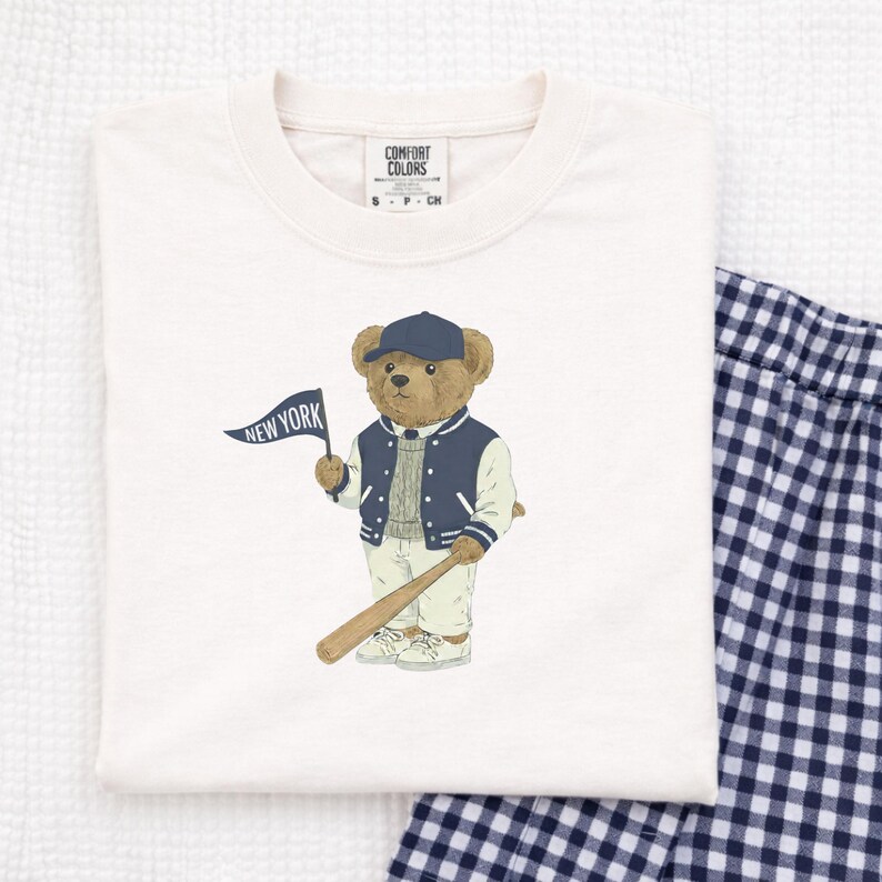Op de afbeelding: Een off-white t-shirt met ronde hals en een afbeelding van een bruine teddybeer met een marineblauwe baseballpet, een marineblauw-wit varsity-jack en een grijze trui. De beer houdt een marineblauwe vlag vast met de tekst NEW YORK en een honkbalknuppel.