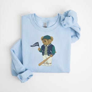 May include: Pale blue sweatshirt with a cartoon bear wearing a baseball cap and jacket, holding a bat and a flag that says "SEATTLE". The bear is brown, and the jacket is navy and green. The sweatshirt is folded.