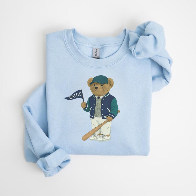 May include: Pale blue sweatshirt with a cartoon bear wearing a baseball cap and jacket, holding a bat and a flag that says "SEATTLE". The bear is brown, and the jacket is navy and green. The sweatshirt is folded.
