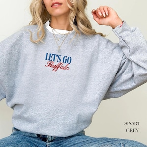 May include: A light gray sweatshirt with the text "LET'S GO Buffalo" in blue and red. The sweatshirt is a crew neck style and has long sleeves. The bottom right corner of the image has the text "SPORT GREY".