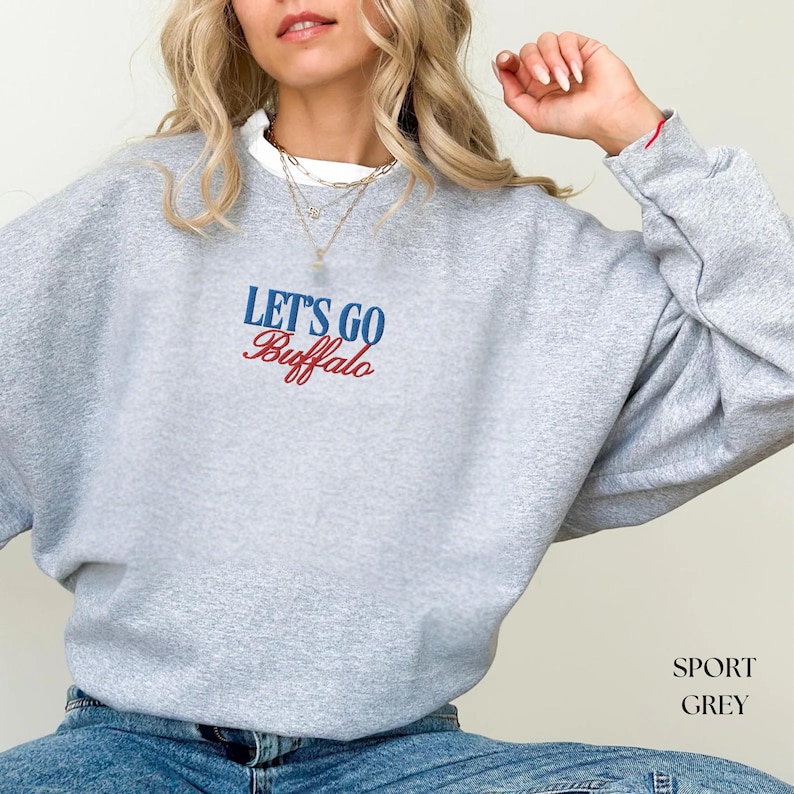 May include: A light gray sweatshirt with the text "LET'S GO Buffalo" in blue and red. The sweatshirt is a crew neck style and has long sleeves. The bottom right corner of the image has the text "SPORT GREY".
