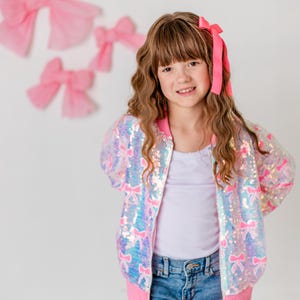 May include: A young person wearing a pink and iridescent sequined bomber jacket with bow accents, a white top, and blue jeans. A pink bow adorns their hair. Pink bows decorate the white background.