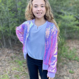 May include: A sequined bomber jacket in shades of pink and purple, with a light purple trim and zipper. The jacket is paired with a light blue shirt and black pants. The outfit is suitable for children.