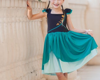 May include: A young girl wearing a blue and green princess dress with a gold leaf design. She is wearing a tiara and has her hair in braids. She is standing in front of a white stone wall.