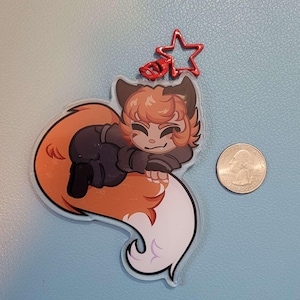 May include: A clear acrylic charm featuring a cartoon fox character with orange and brown fur, wearing a black outfit. The charm has a red star-shaped clasp. A U.S. quarter is next to the charm for size comparison.