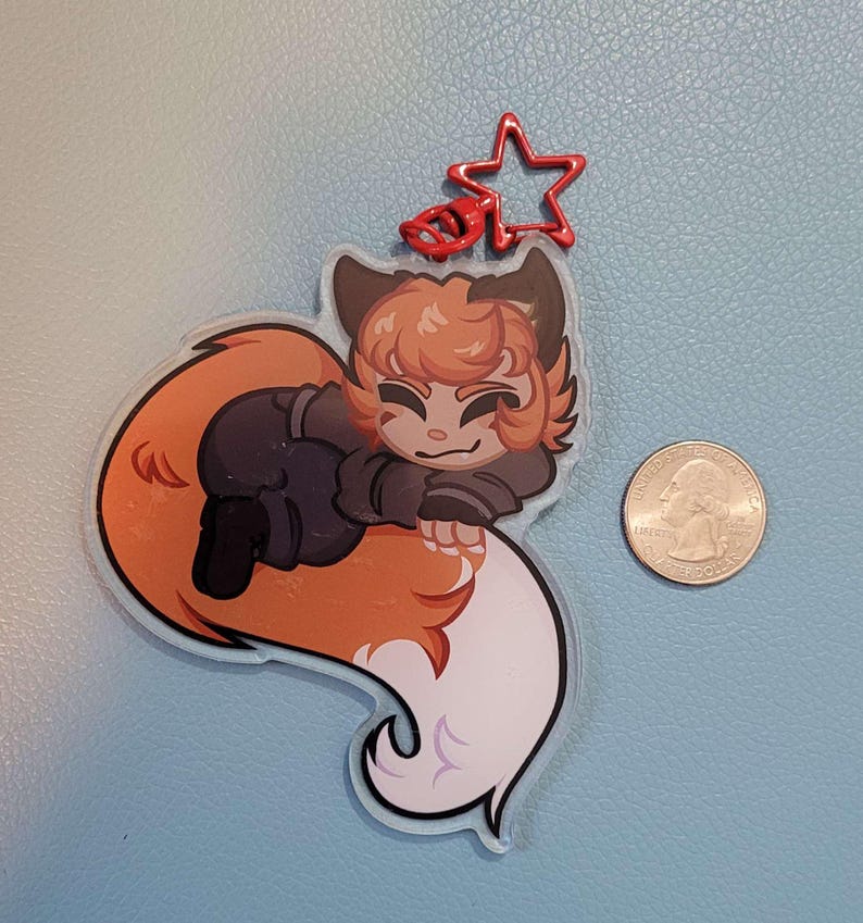May include: A clear acrylic charm featuring a cartoon fox character with orange and brown fur, wearing a black outfit. The charm has a red star-shaped clasp. A U.S. quarter is next to the charm for size comparison.