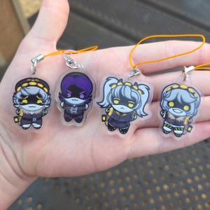 May include: Four clear acrylic charms featuring stylized cartoon characters with yellow eyes and dark outfits. Each charm includes a small metal loop and an orange string for hanging. The characters have distinct hairstyles and expressions, with the text ENDOFTHEVERSE printed on each piece.