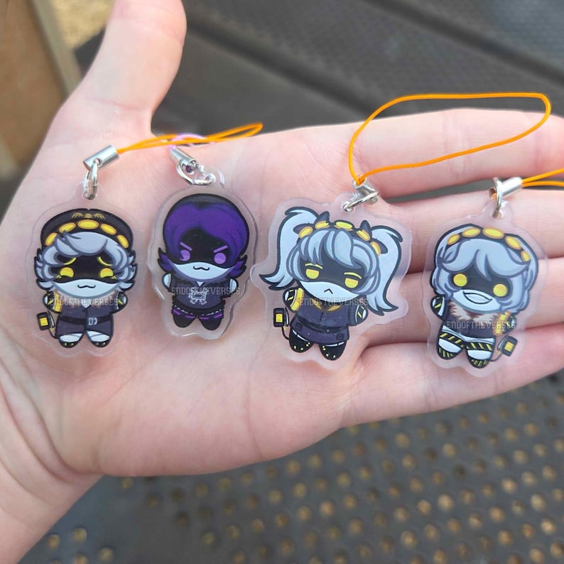 May include: Four clear acrylic charms featuring stylized cartoon characters with yellow eyes and dark outfits. Each charm includes a small metal loop and an orange string for hanging. The characters have distinct hairstyles and expressions, with the text ENDOFTHEVERSE printed on each piece.
