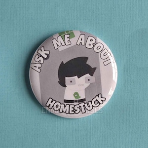 May include: A circular pinback button featuring a cartoon character with black hair and glasses, wearing a white shirt with a small green ghost graphic. The text ASK ME ABOUT HOMESTUCK is printed in black capital letters around the character.