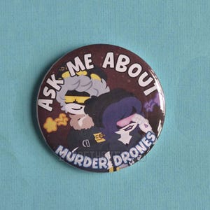 May include: A circular pin-back button featuring a dark purple background with two animated characters. One character has grey hair and yellow visor glasses, while the other has dark purple hair and a pink visor. The text reads ASK ME ABOUT MURDER DRONES.