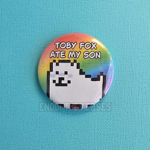 Puede incluir: Una chapa redonda con una ilustración pixelada de un perro blanco sobre un fondo de degradado arcoíris. El texto "TOBY FOX ATE MY SON" está impreso en letras mayúsculas negras y negritas sobre la ilustración del perro en la superficie.