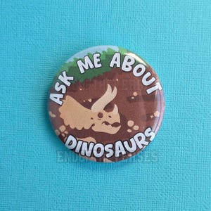 Puede incluir: Una chapa redonda con la ilustración de un triceratops marrón sobre un fondo de tierra marrón y un borde superior verde que representa hierba. El texto ASK ME ABOUT DINOSAURS está impreso en letras mayúsculas blancas a lo largo del borde.