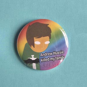 May include: A circular pinback button featuring a rainbow gradient background and a cartoon character with brown hair and white eyes. The character wears a green shirt with a white cross symbol. Text reads: Andrew Hussie killed my family.