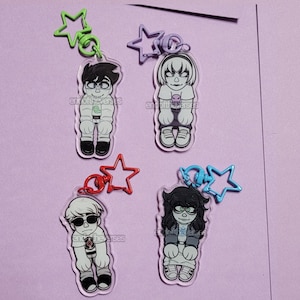 May include: Four acrylic keychains featuring cartoon characters. Each keychain has a different character design, with black and white outfits and star-shaped attachments in green, purple, red, and blue. The keychains are on a purple background.