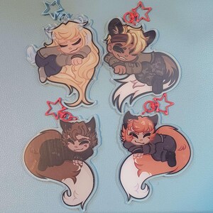 May include: Four acrylic charms featuring cartoon characters with animal-like features. The characters have various hair colors and are wearing different outfits. Each charm has a star-shaped clasp in either blue or red.