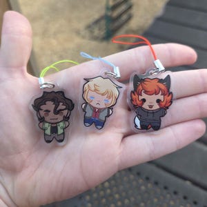 May include: Three clear acrylic charms featuring cartoon characters with different hair colors and outfits. Each charm has a colored lanyard attached. The characters include a brown-haired figure in a green jacket, a blonde figure in a jacket, and a red-haired figure with fox ears.